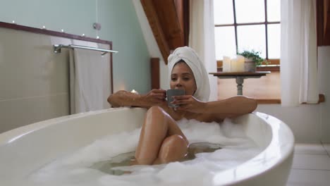 mixed race woman wearing towel on head taking a bath and drinking coffee