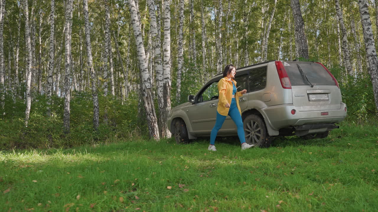Campeggiatrice che scarica l'attrezzatura dal bagagliaio di un SUV, donna con giacca gialla che solleva un frigo portatile e casse, organizza le scorte sull'erba accanto a betulle, preparazione pratica per l'allestimento del campeggio, movimento concentrato.