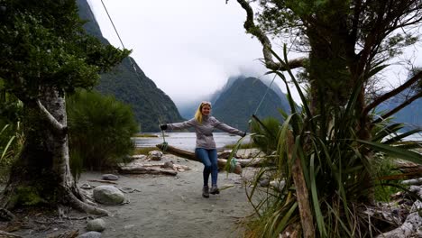 Blonde-white-woman-is-sitting-on-a-swing-in-Milford-Sound-with-a-stunning-background,-while-smiling-and-looking-in-the-sky-between-trees