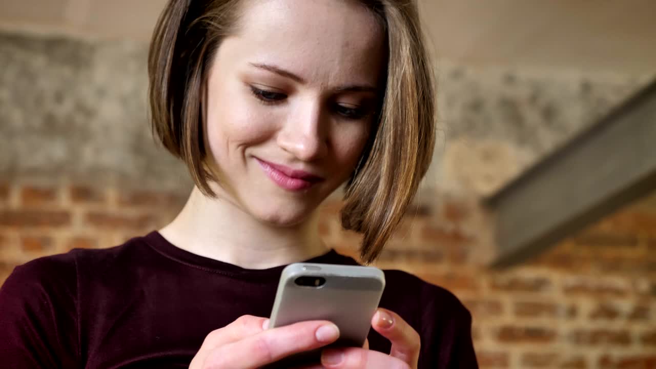Giovane e bella ragazza sta digitando un messaggio sul suo smartphone, sorridendo, concetto di comunicazione, sfondo in mattoni