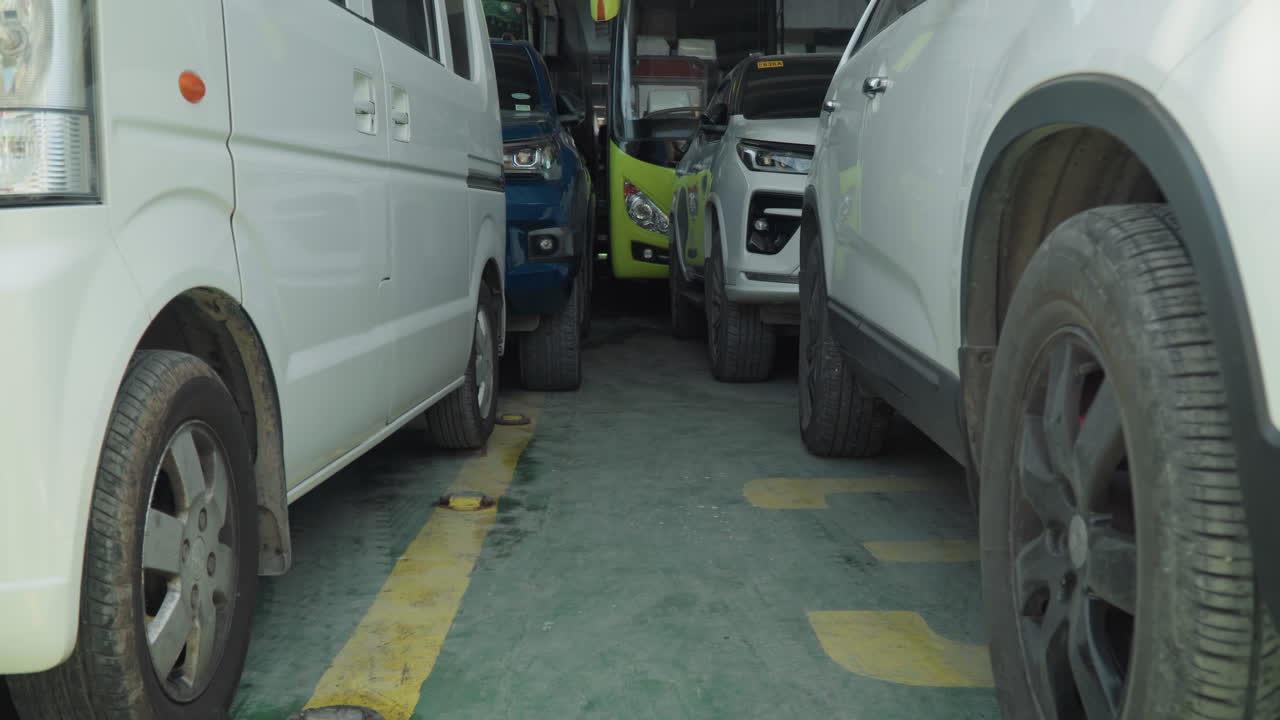 Estacionamiento urbano apretado entre furgonetas blancas, primer plano de ruedas y neumáticos, asfalto agrietado con marcas de carril amarillas, espacio estrecho entre vehículos, manchas de aceite y suciedad, ambiente de operación de entrega tensa, atardecer.