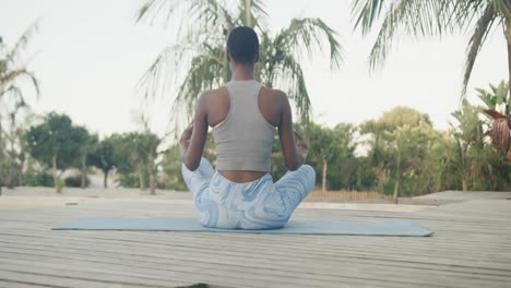 Rear-view-of-biracial-woman-practicing-yoga-meditation-sitting-on-jetty-by-palm-trees,-slow-motion