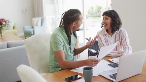 mujer biracial feliz en silla de ruedas y compañero masculino sonriente con papeleo y computadora portátil en la sala de estar