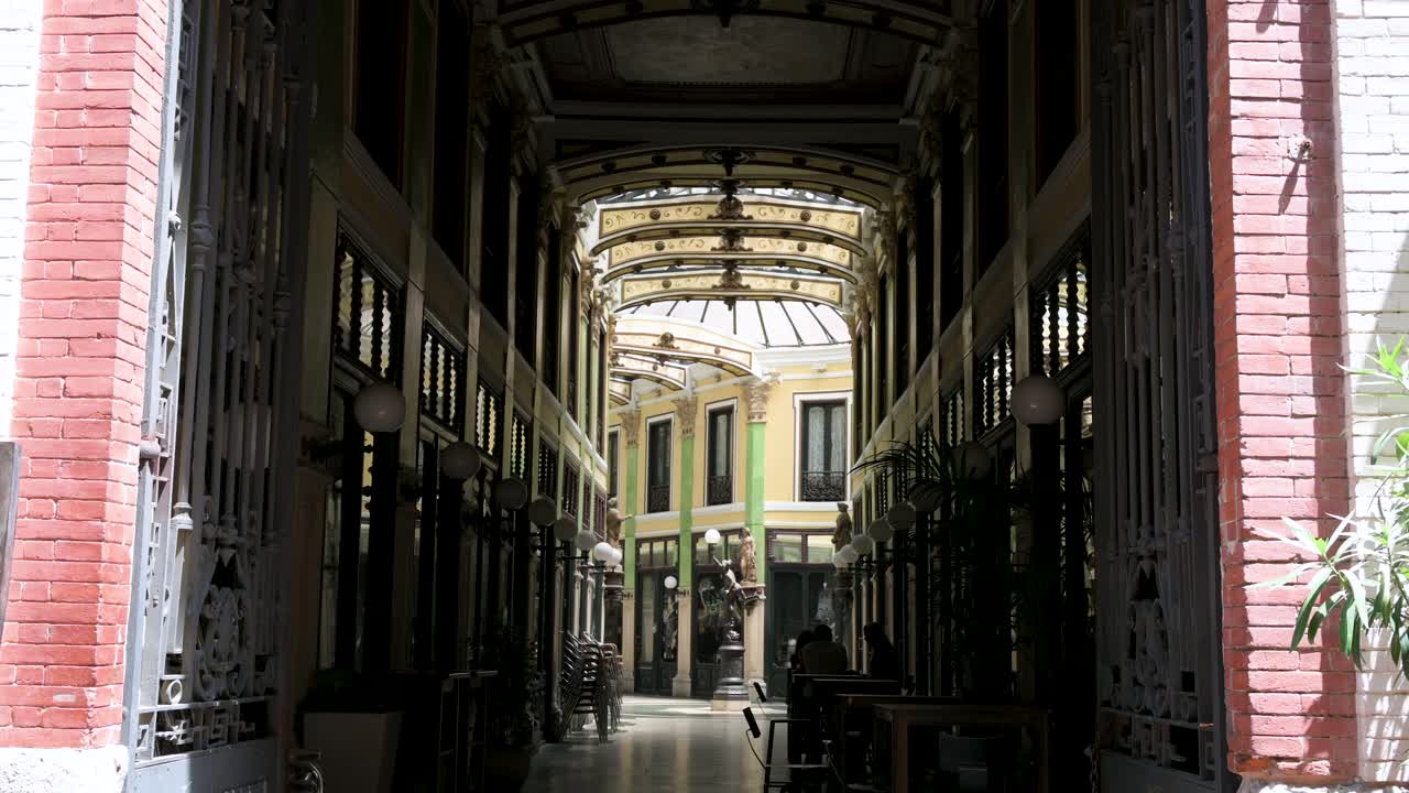 Tilt-down entrance view of Pasaje Gutiérrez in Valladolid, a 19th-century covered gallery with a glass roof, showcasing unique architecture and shops that blend history and modern commerce in Spain.