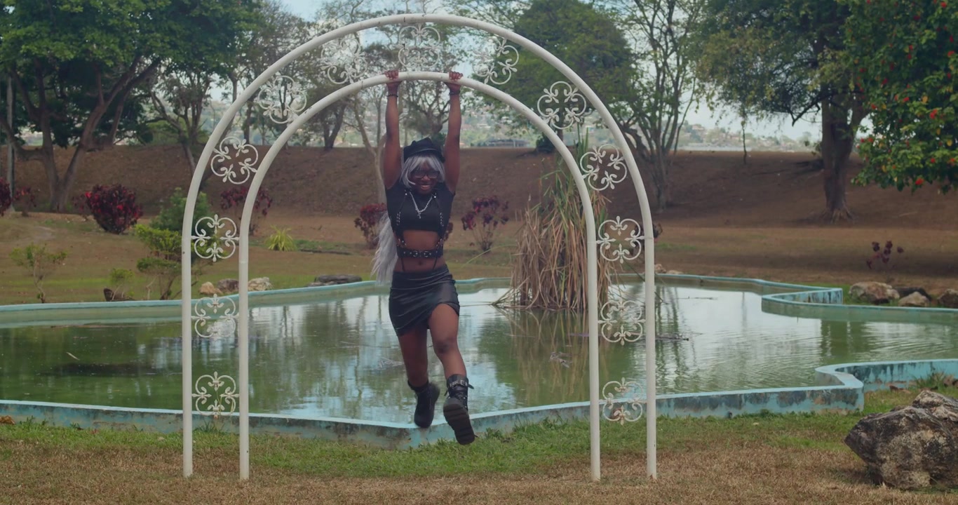 Close-up slow-motion shot of a dark-skinned woman in a stylish leather outfit and boots looking into the camera at a vibrant Caribbean park under a brilliant sun.