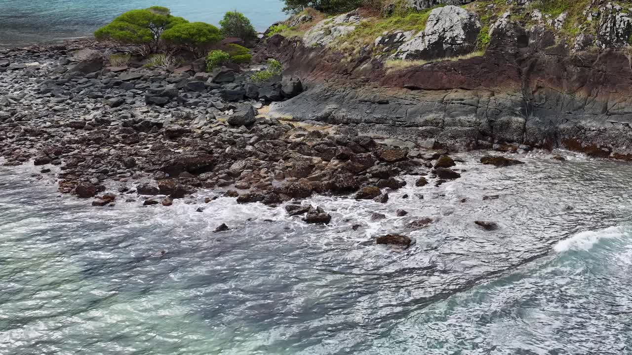 Aerial drone footage pulls back from a rocky, vegetated coastline to reveal waves meeting the shore and the expansive Coral Sea under natural daylight
