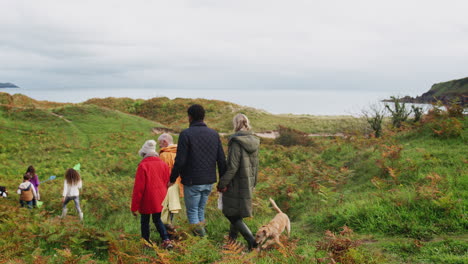 Vista-Trasera-De-Una-Familia-Activa-Multigeneracional-Con-Un-Perro-Caminando-Por-El-Sendero-Costero-De-Otoño