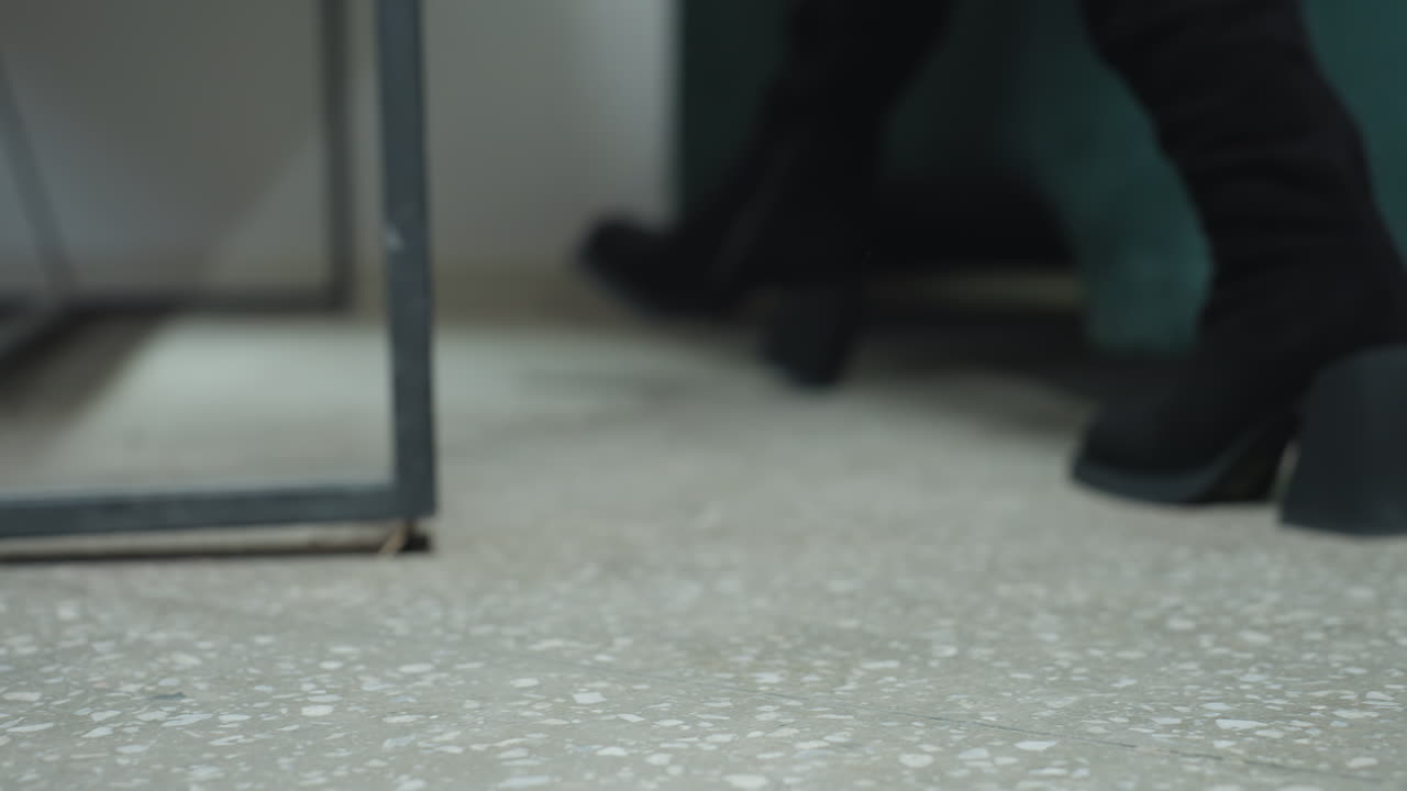 Low angle close up shows woman in stylish black heel boots stepping across polished terrazzo floor toward green couch, preparing to sit in modern professional workspace environment