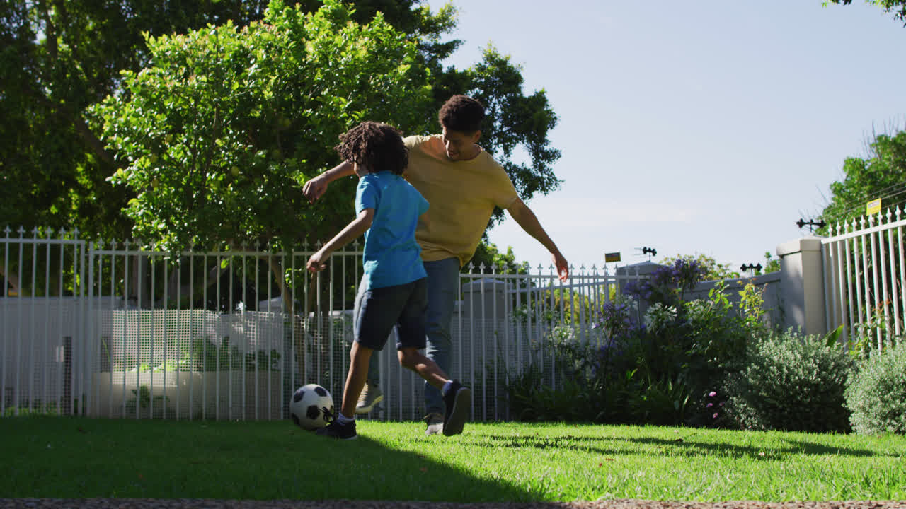feliz hombre biracial y su hijo jugando al fútbol en el jardín