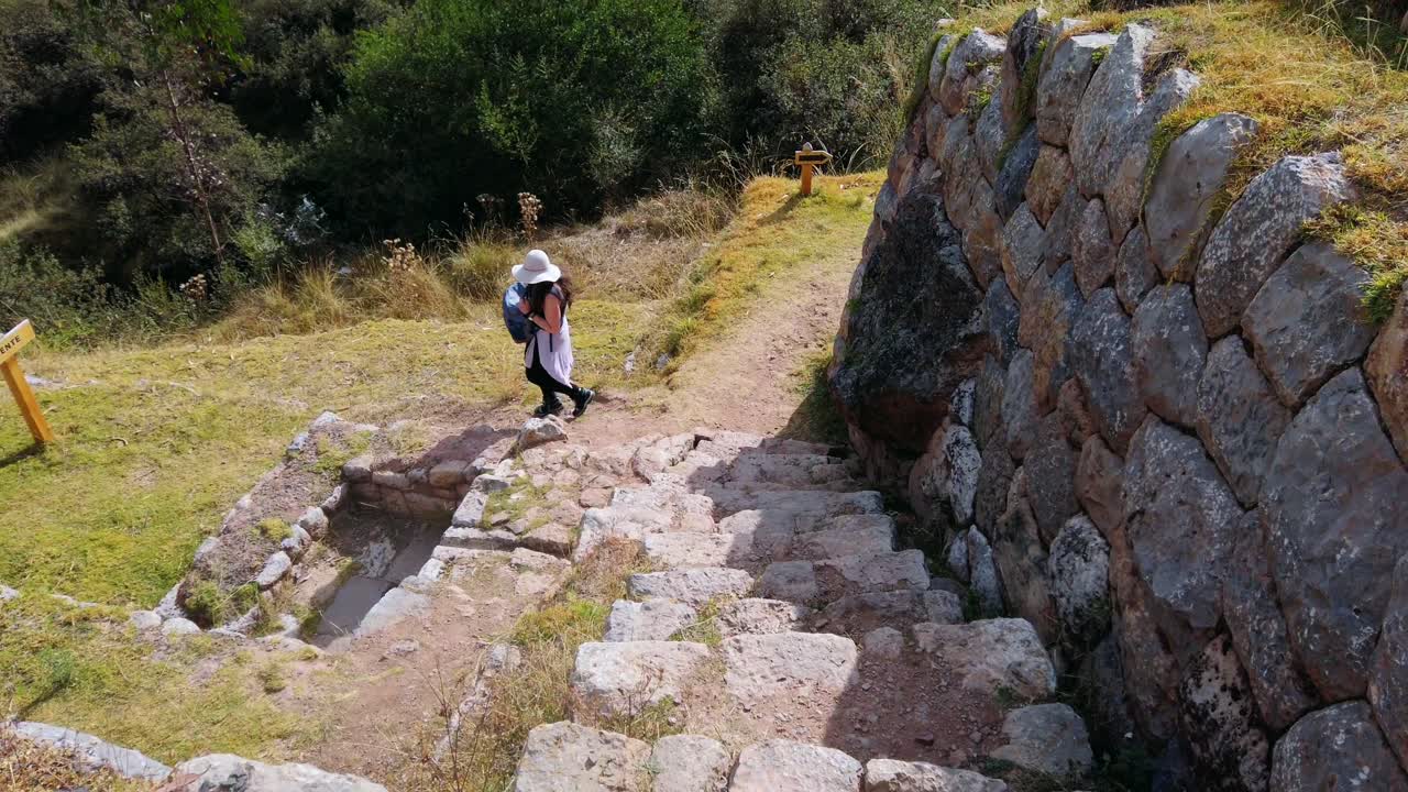 Young brunette tourist walking through the ancient ruins of Inkilltambo, wearing a Peruvian-style hat and carrying a backpack. Solo traveler exploring historic Inca site