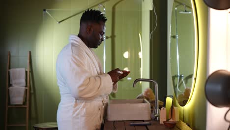 side view positive handsome african american man in bathrobe turning on music on smartphone applying shave cream. portrait of confident young guy shaving in bathroom at home in the morning.