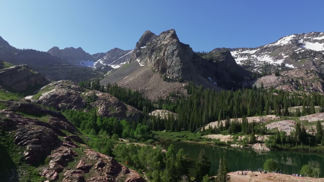 Ripresa aerea con drone che zooma lentamente sul lago Blanche nel Big Cottonwood Canyon vicino a Salt Lake City, nello Utah, in una luminosa mattina estiva e verdeggiante con cime rocciose, vegetazione lussureggiante e il Sundial Peak che svetta in alto