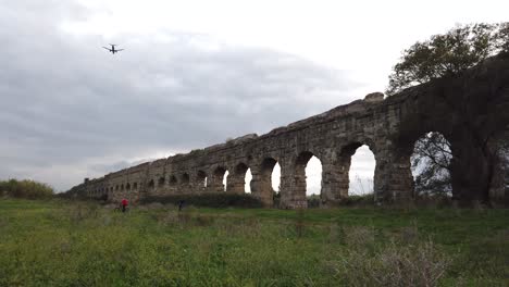 Commercial-Plane-flying-over-an-aqueduct-from-ancient-Rome-in-parco-degli-acquedotti-in-the-outskirts-of-the-capital-of-Italy,-tilt-down-move-for-reveal-and-wide-angle