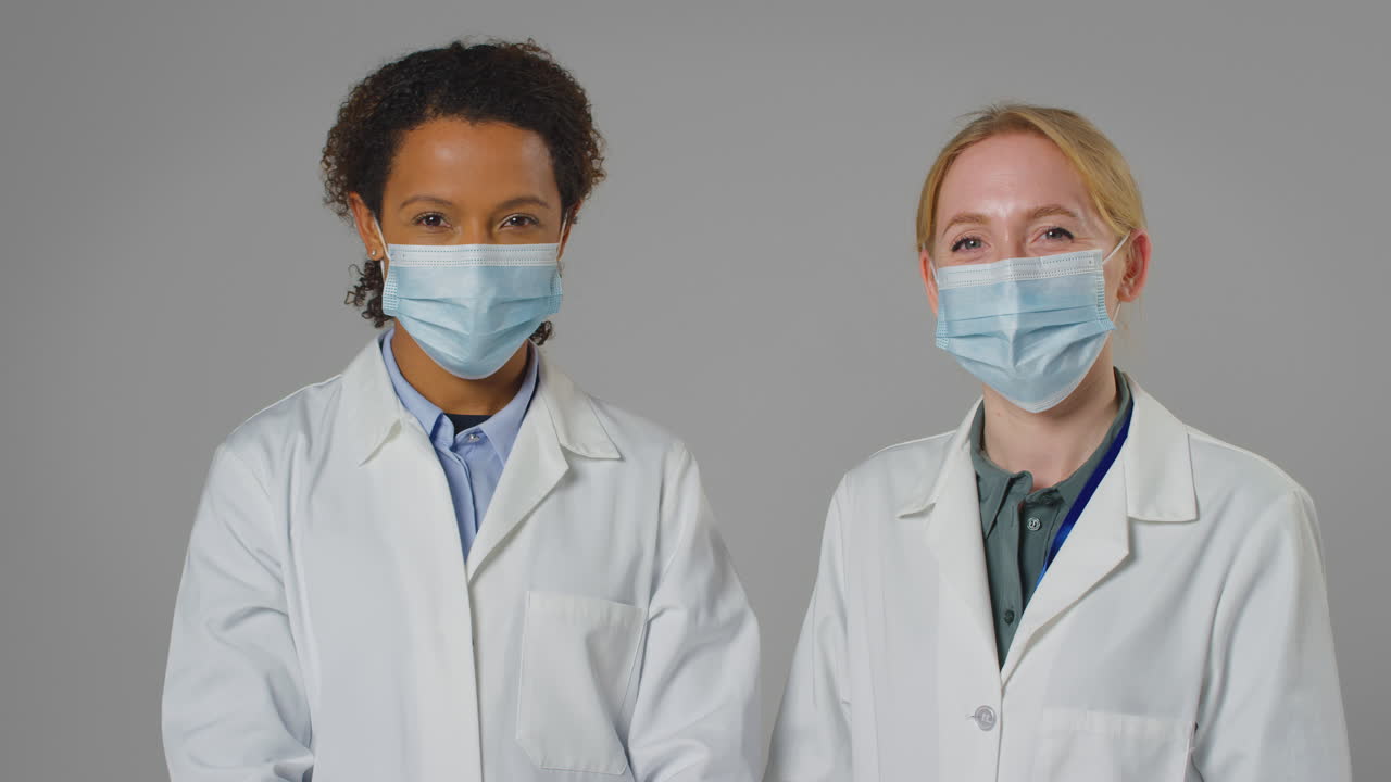 Studio Portrait Of Two Female Doctors Or Lab Workers Wearing Face Masks In White Coats