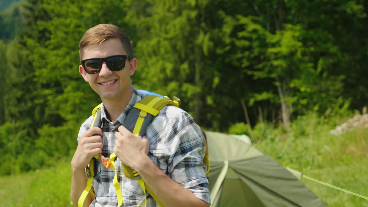 un joven con una mochila está mirando la cámara de pie en el fondo de una tienda de campaña po