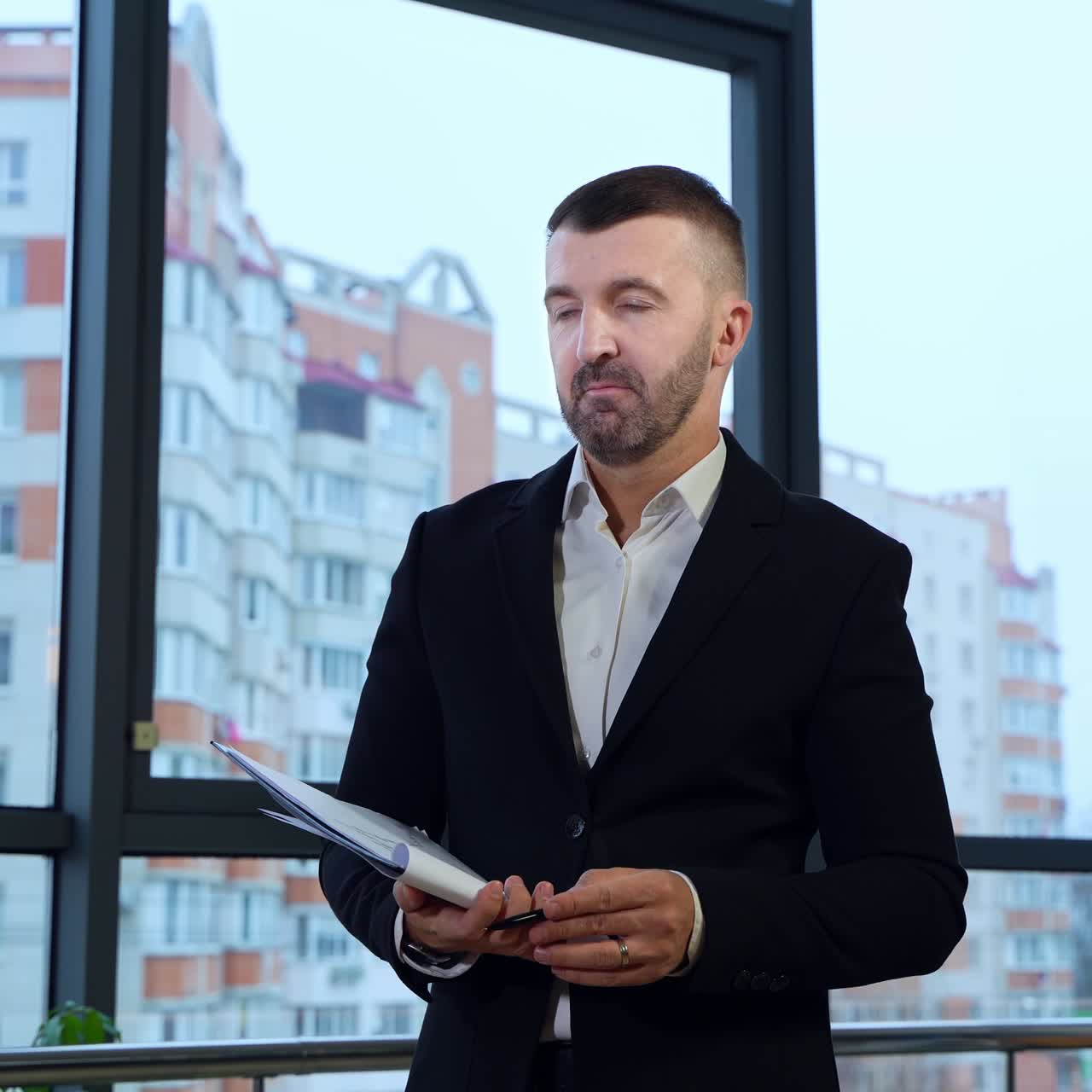 Middle-aged boss in suit standing in front of employees and holding papers. Man replying questions of his staff against city backdrop