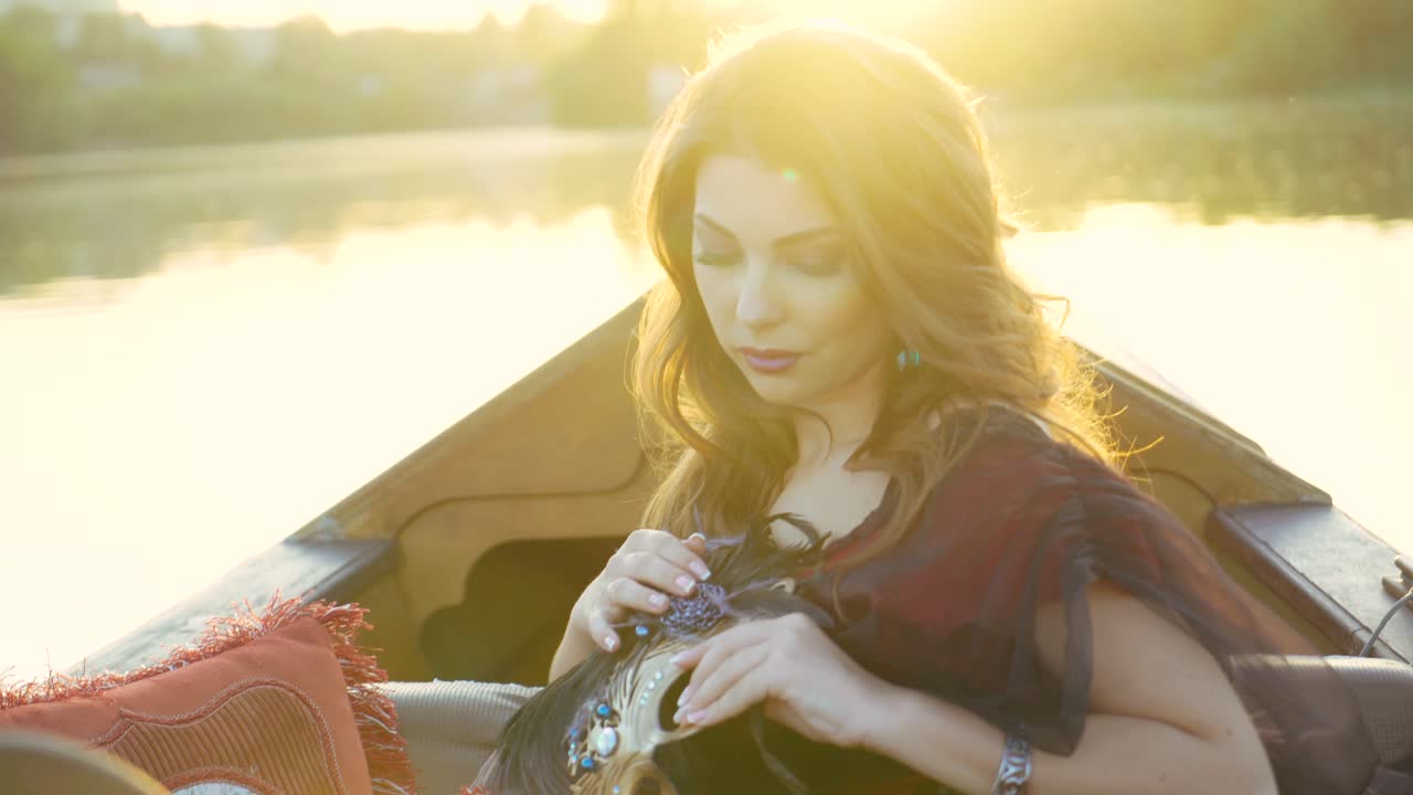 A beautiful woman is holding a luxurious carnival mask in her hands. The lady examines it in the gondola on the background of sunset. Close-up