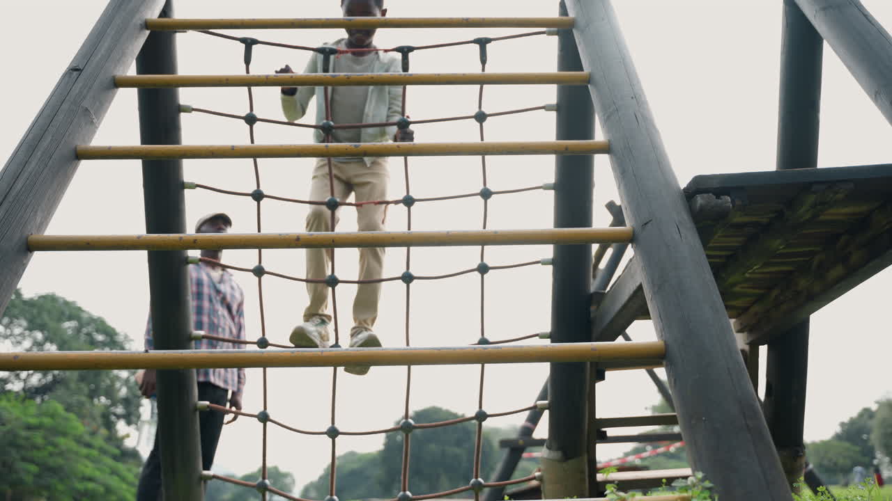 Black father coaching son on ladder rungs in park training session, dad spotting from side, balance practice across vertical rope ladder, focused steps, supportive instruction and supervision