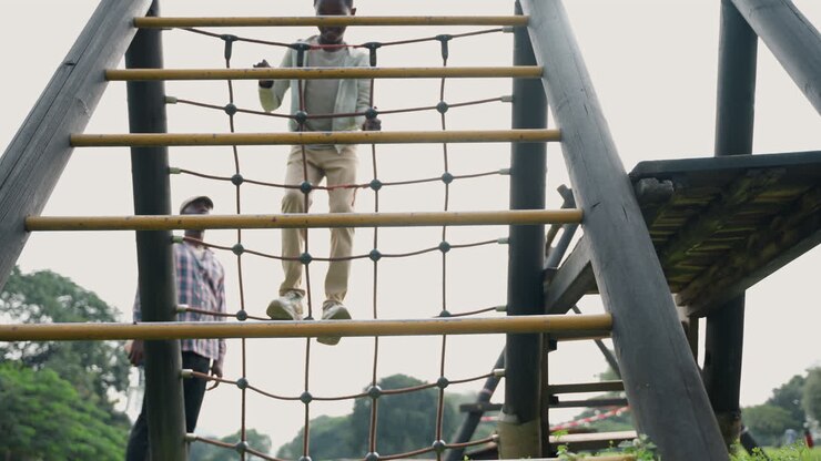 Black father coaching son on ladder rungs in park training session, dad spotting from side, balance practice across vertical rope ladder, focused steps, supportive instruction and supervision