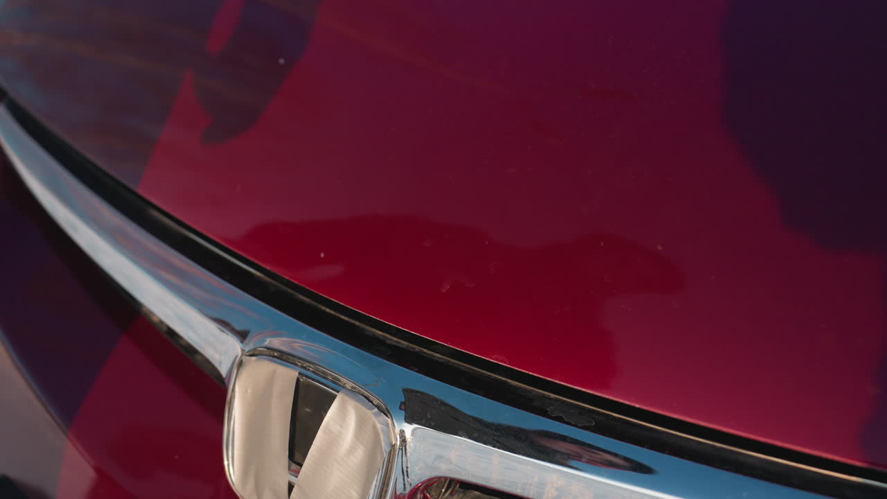 Close-up of person's hand pressing down on slightly open red car bonnet to shut it, with sunlight casting clear reflection of individual on glossy hood during cold day in snowy outdoor environment