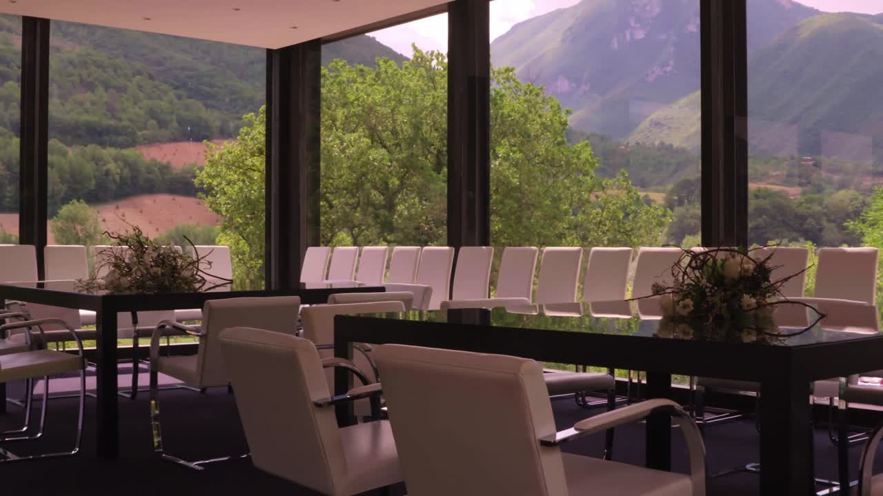 A wide shot pans left, showcasing a modern dining area with white chairs, floral centerpieces, and large windows framing a stunning view of rolling hills and mountains.