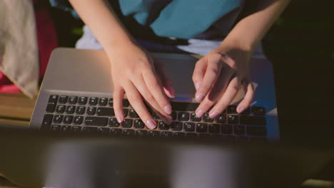 close-up of woman seated outdoors at night, typing on laptop with hands illuminated by screen glow, denim jeans reflect subtly on laptop surface, with a red bag nearby