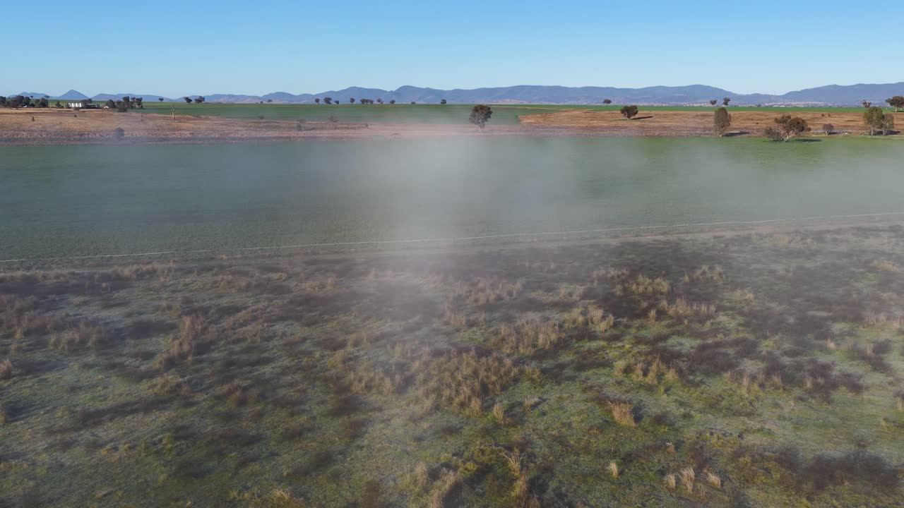 Drone glides above a foggy meadow with distant mountains, early morning sunlight, and soft mist creating a tranquil, natural landscape scene