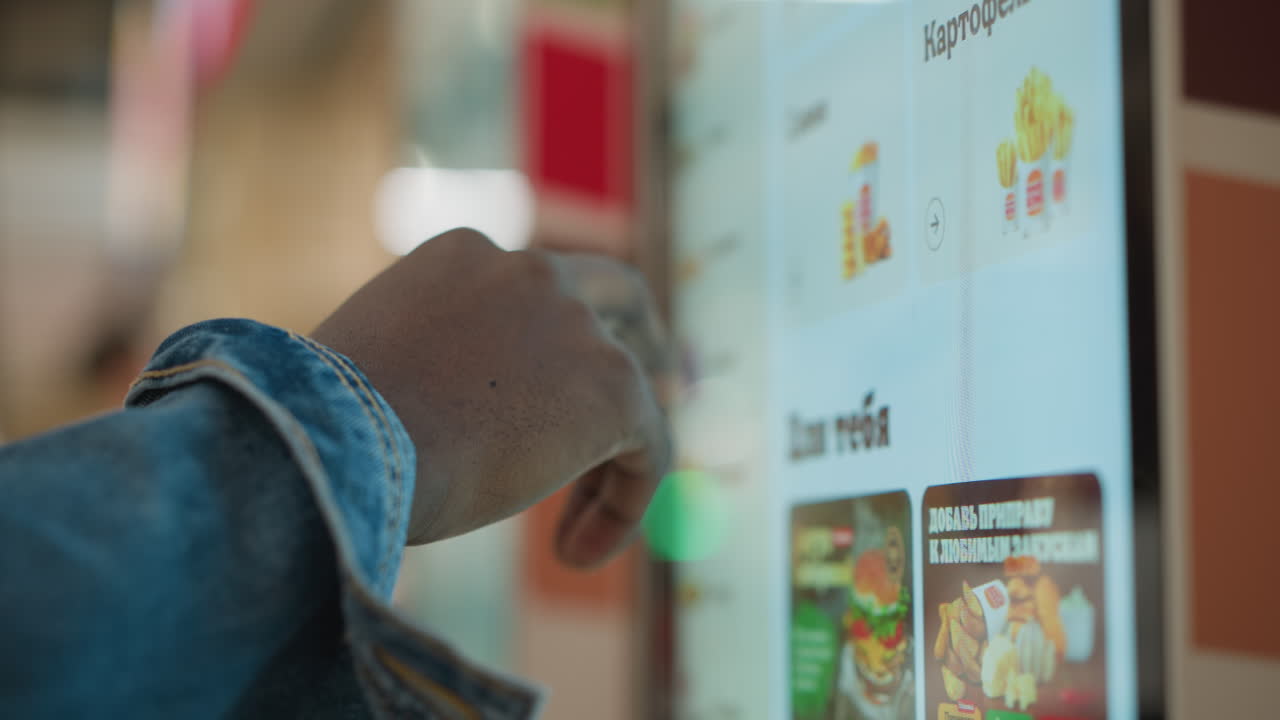 Close-up of hand with denim sleeve pointing at digital menu screen displaying food options, including fries, in a modern fast-food setting