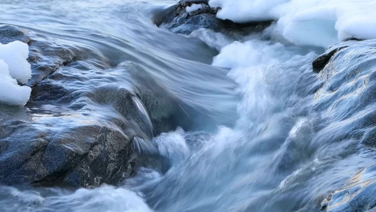 Vista ravvicinata di acqua che scorre velocemente e si precipita diagonalmente attraverso un letto di ruscello roccioso, con chiazze di neve e ghiaccio lungo le rive sotto una fresca luce naturale.