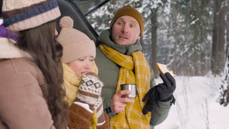 Side-View-Of-Parents-And-Daughter-Eating-Sandwiches-And-Drinking-Hot-Drink-Sitting-In-The-Trunk-Of-The-Car-In-A-Snowy-Forest