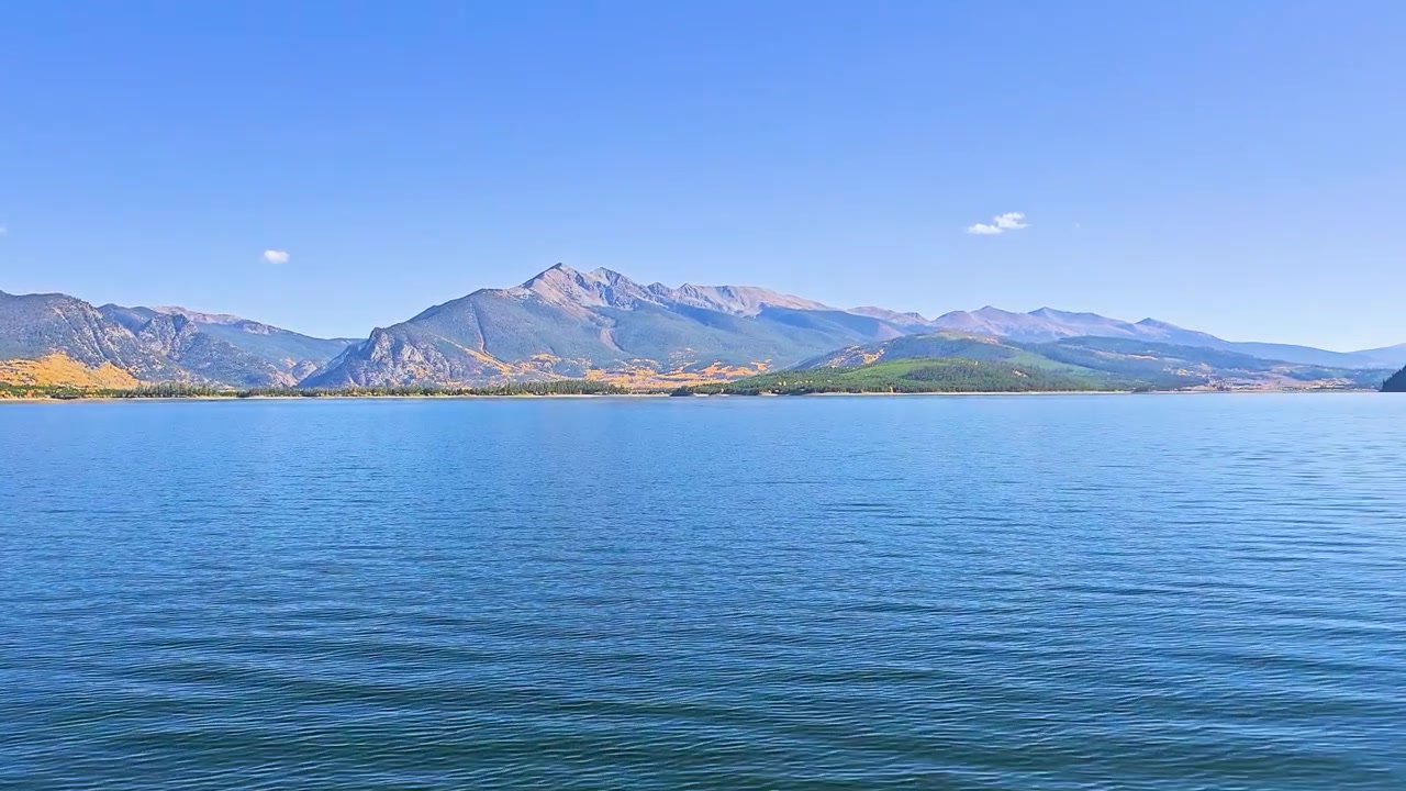 動くボートから見た前方に向かう湖の眺め。さざ波立つ水面、なだらかな山々、黄色い秋の色が点在する松に覆われた斜面、そして白い雲が浮かぶ広々とした青空が広がっています。