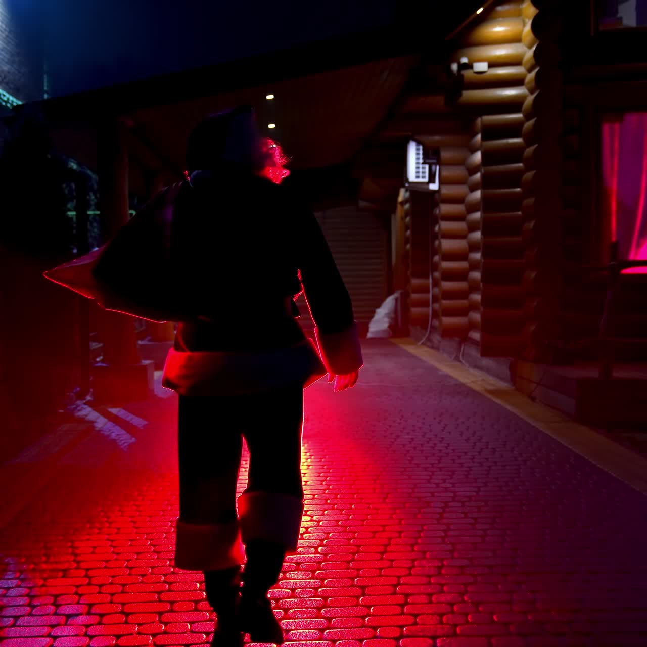 Santa Claus with bag walks on street at Christmas night. Silhouette of Santa carrying presents for children in winter at night. Christmas tree in window. Back view