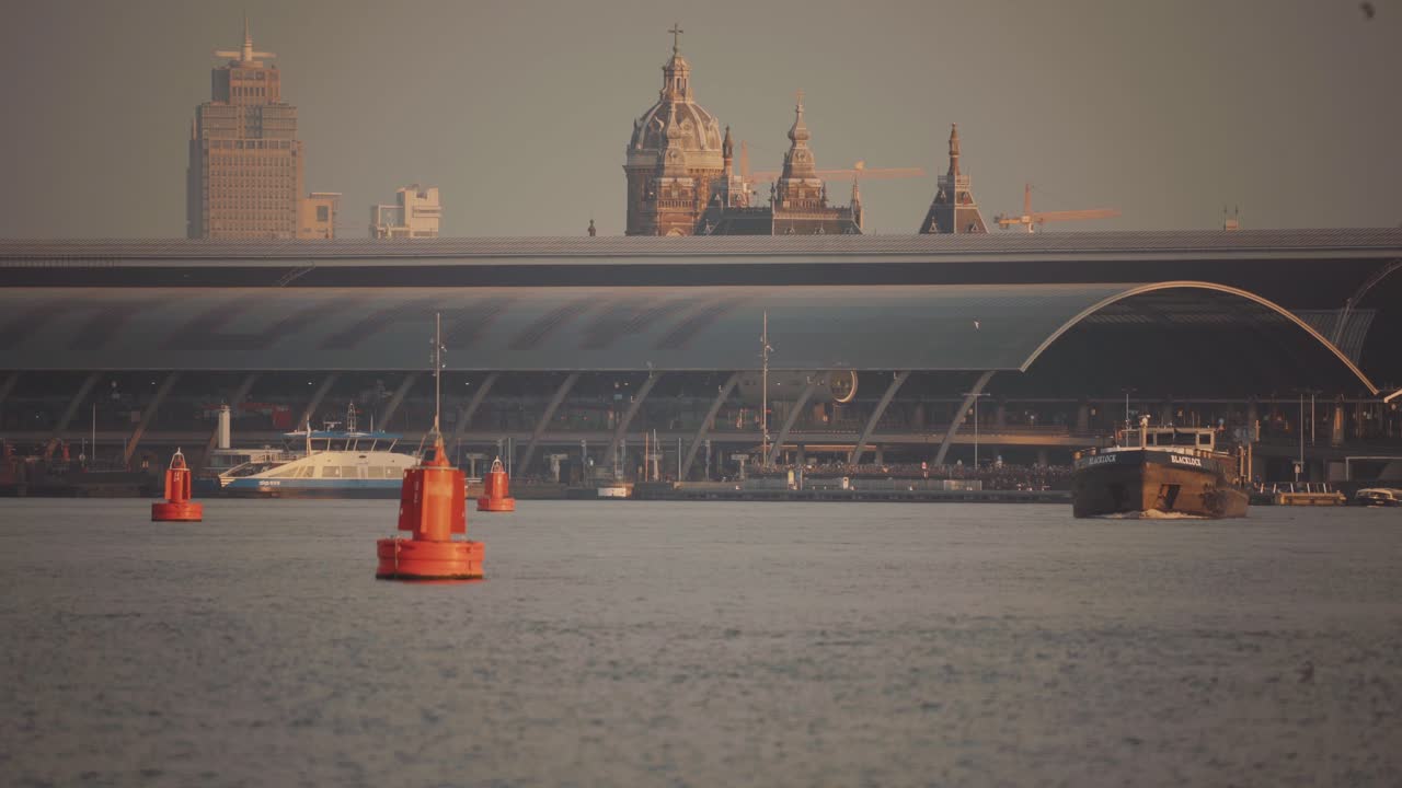 Boats floating through ij river with Amsterdams central train station and skyline behind it, telephoto lens