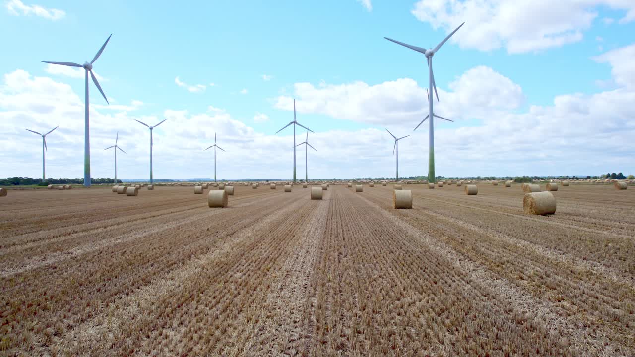 Captured from above, a row of wind turbines gracefully turns in a Lincolnshire farmer's freshly harvested field, with golden hay bales in the foreground