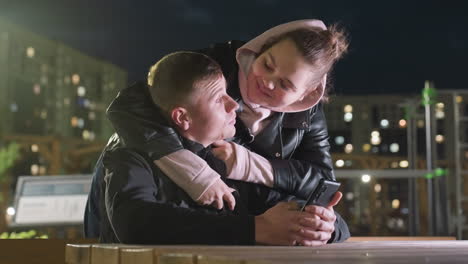 professional man seated operating phone at night in city as friend walks up behind him and places hands on his shoulder in warm gesture under streetlight with blurred urban background