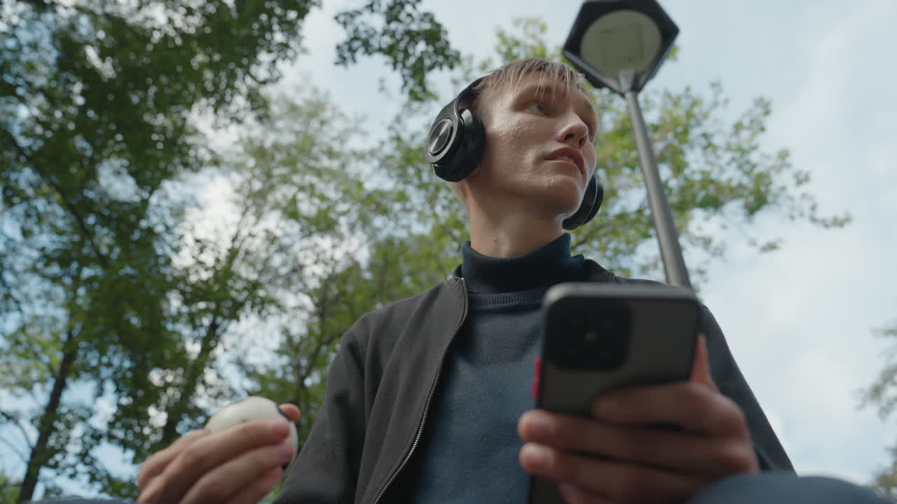 Giovane uomo caucasico con cuffie che scorre sullo smartphone e tiene in mano una pallina sotto un lampione in un parco alberato, ritratto candido dal basso, maglione casual, cuffie over-ear, espressione pensierosa, nuvoloso