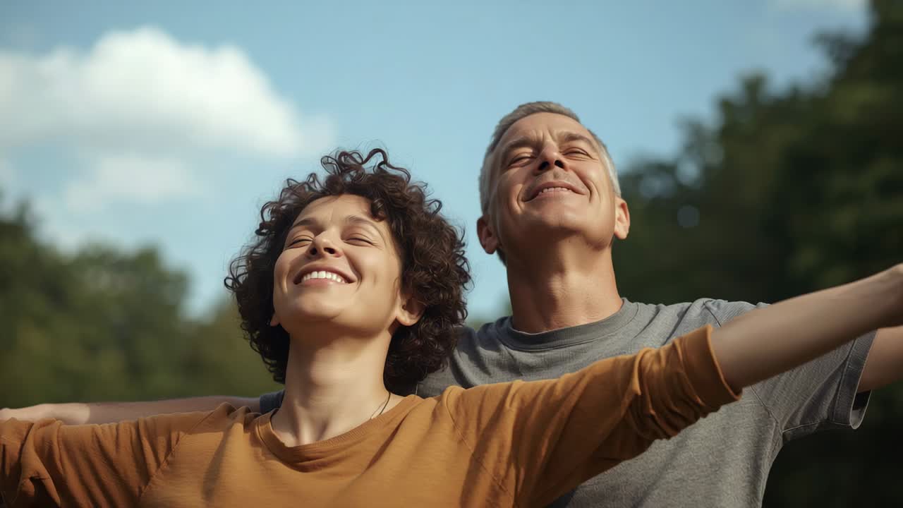 Casal de braços abertos, inclinando a cabeça para trás, com o sol aquecendo seus rostos no parque, exibindo blusa ferrugem e colar