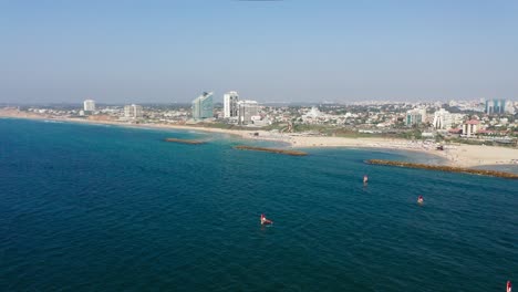 Mehrere-Windsurfer-Surfen-Auf-Dem-Klaren-Blauen-Wasser-Des-Mittelmeers-In-Richtung-Der-Wellenbrecher-Mit-Im-Hintergrund-Der-Touristischen-Stadt-Herzeliya-In-Israel