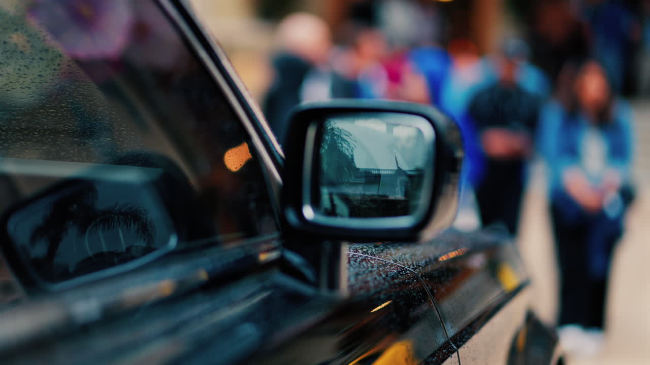 Close up of the mirror of a black car in the rain with people walking on a blurred background