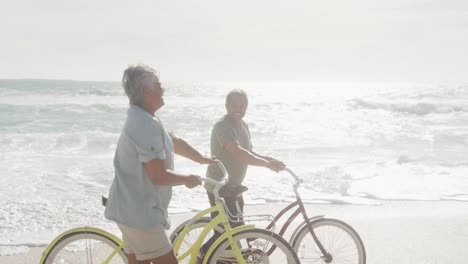 side view of happy hispanic senior couple walking with bikes on beach at sunset