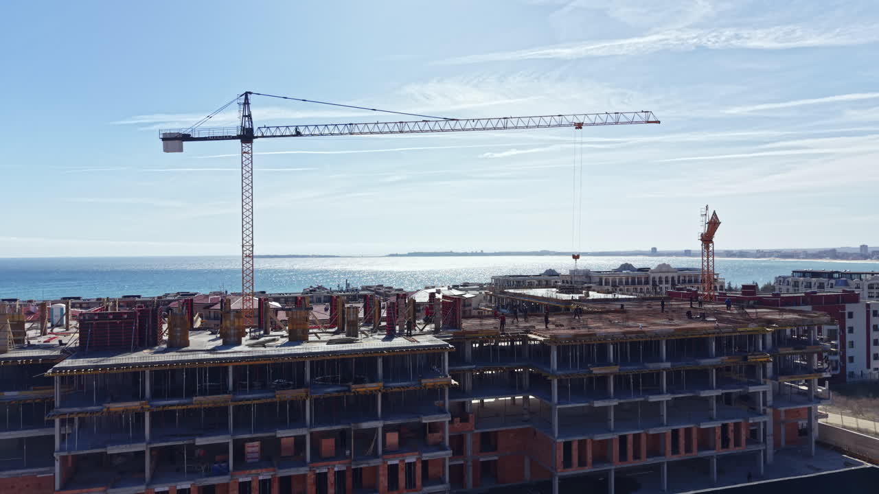 Lavori di costruzione su un edificio vicino all'oceano con una gru e cielo sereno, osservati da un punto elevato durante il giorno.