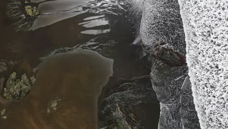 aerial drone flying over black sandy secluded beach with white waves crashing onto shore