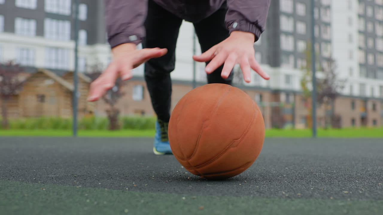 Seseorang melakukan push-up dengan bola basket di jalanan lapangan, seorang atlet melatih kekuatan inti menggunakan bola basket sebagai penyangga di lapangan aspal basah dekat gedung apartemen di bawah langit mendung, mengenakan beanie dan jaket