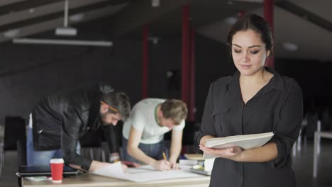 Portrait-of-a-fashionable-young-professional-woman-holding-folders-with-papers-and-taking-notes-in-a-stylish-contemporary-office.-Office-workers-on-the-background.-Shot-in-4k