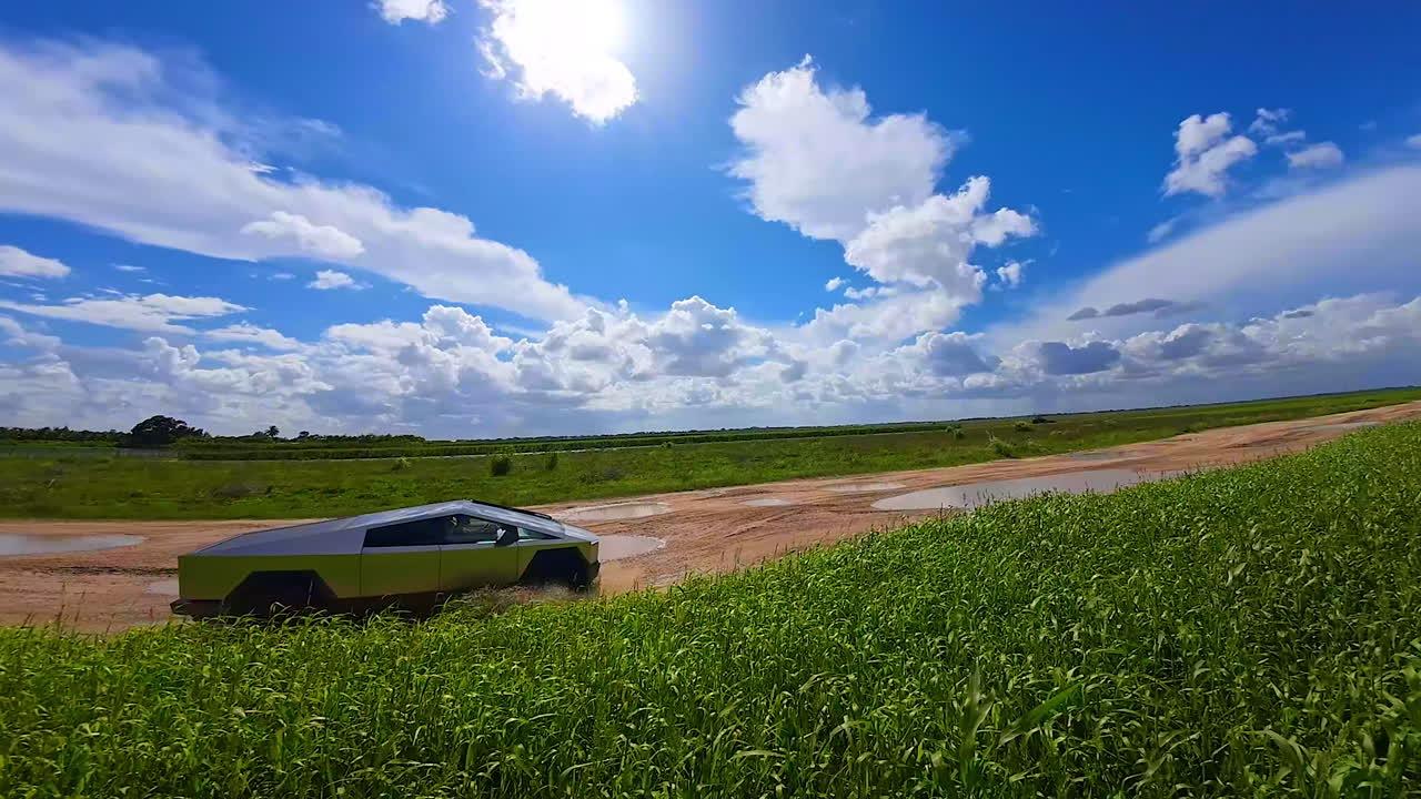 Tesla Cybertruck rides by the ground road with multiple puddles. FPV drone footage of a technological car on sunny day.