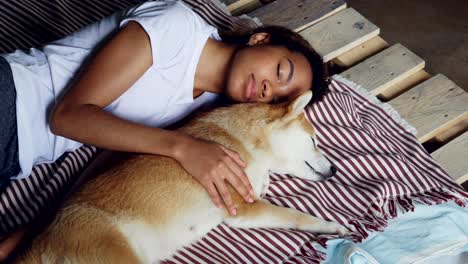una hermosa adolescente afroamericana y un adorable perro mascota están durmiendo juntos en una cama de madera, la chica lleva un cómodo pijama y abraza al animal.