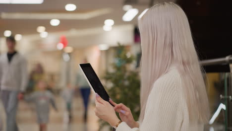 side view urban dweller seated on mall bench tapping tablet screen with manicured nails under bright ceiling lights, blurred crowd browsing stores and strolling through spacious shopping concourse