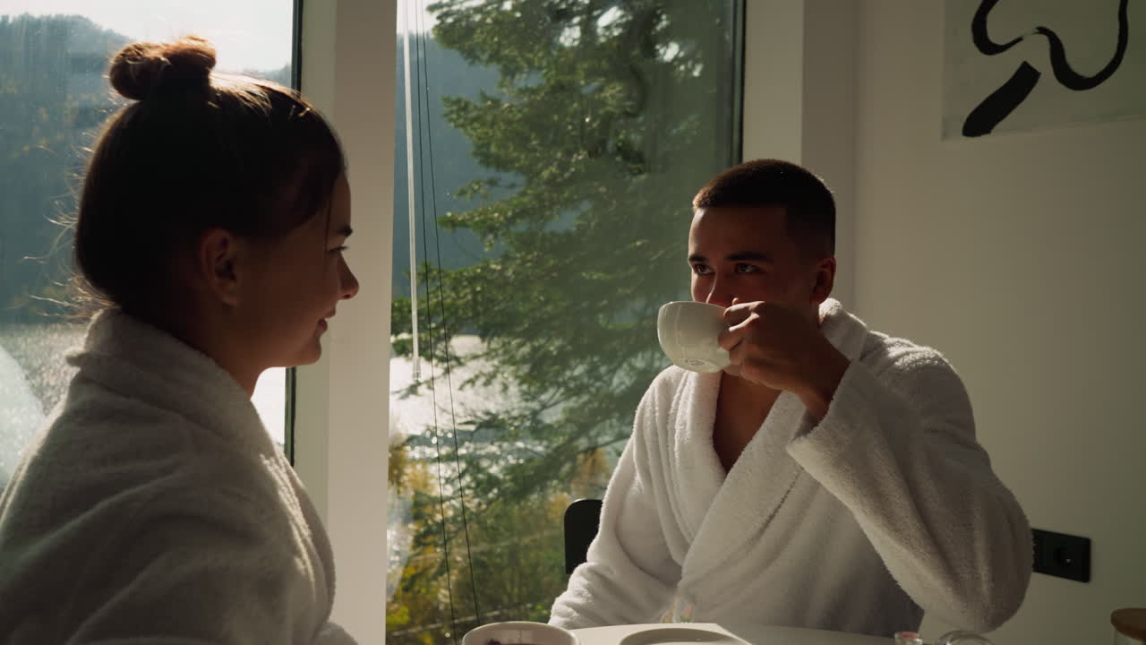 Man drinks tea looking at girlfriend by window. Young couple in terry bathrobes looks happy enjoying breakfast in hotel with picturesque view. Romantic moment