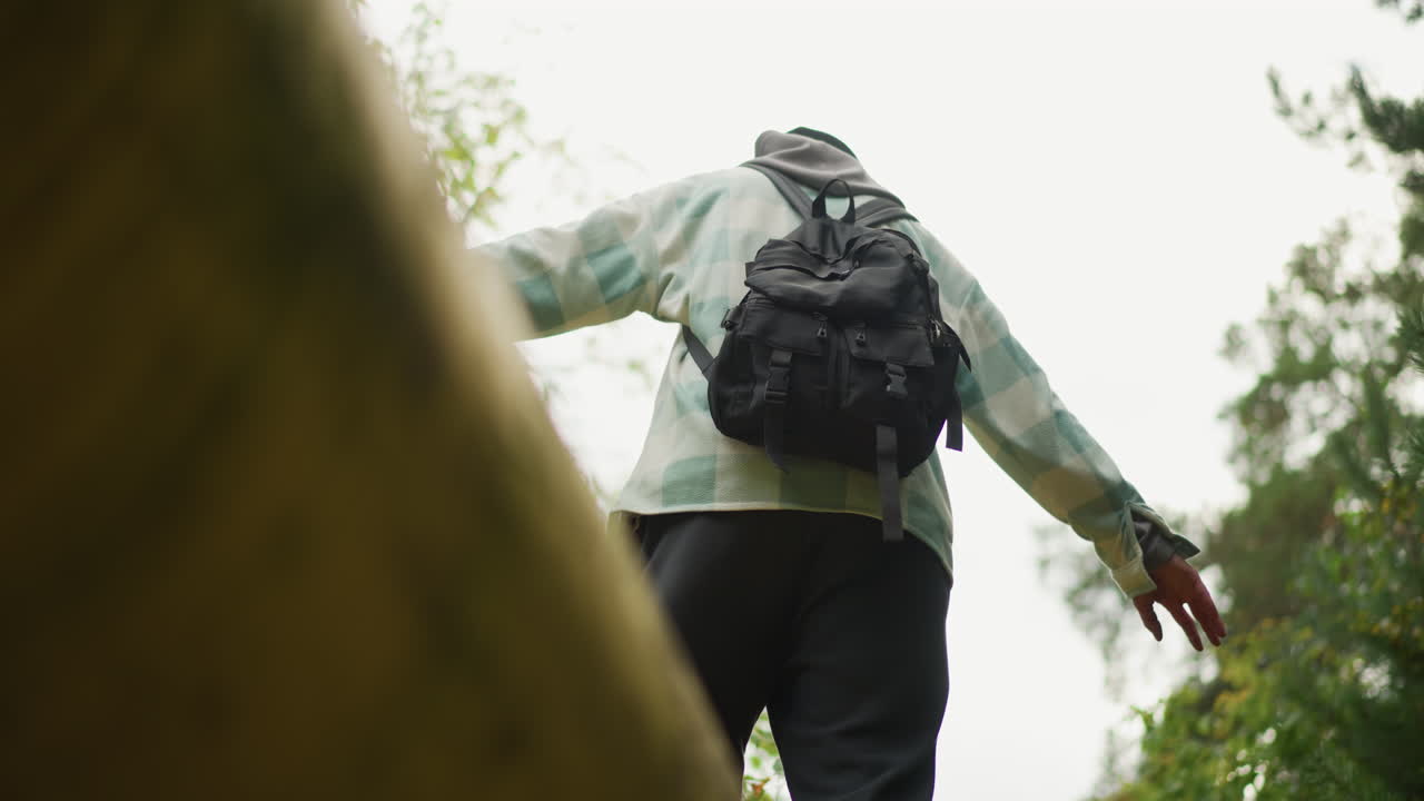 low angle hiker crossing yellow pipeline, dramatic upward perspective of backpacked traveler extending arms for balance with leaves and canopy framing scene, cloudy sky, dynamic motion and triumphant