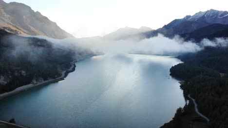 Un-Sobrevuelo-Aéreo-Del-Amanecer-A-Través-De-Las-Nubes-Sobre-El-Lago-Sils-En-Maloja,-Suiza,-Con-Vistas-A-Los-Picos-De-Engadin-Y-Las-Nubes-Moviéndose-Sobre-El-Agua